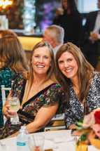 The bride's mom and her friend smile at the wedding reception as they sit down to enjoy their drinks together.