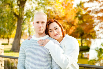 A woman snuggles against a man as they gaze into the camera while standing atop a stone bridge during their fall photoshoot at White Rock Lake in Dallas, TX. Image taken by Photography of Sarah Mae.