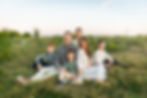 Family of six sitting on a grassy field at sunset, smiling. They're casually dressed in earthy tones, with a serene sky in the background. Image captured by Photography of Sarah Mae.