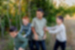 Four boys laughing and playing in a grassy area with trees in the background. They are wearing casual clothes; the mood is joyful and playful. Image captured by Photography of Sarah Mae.