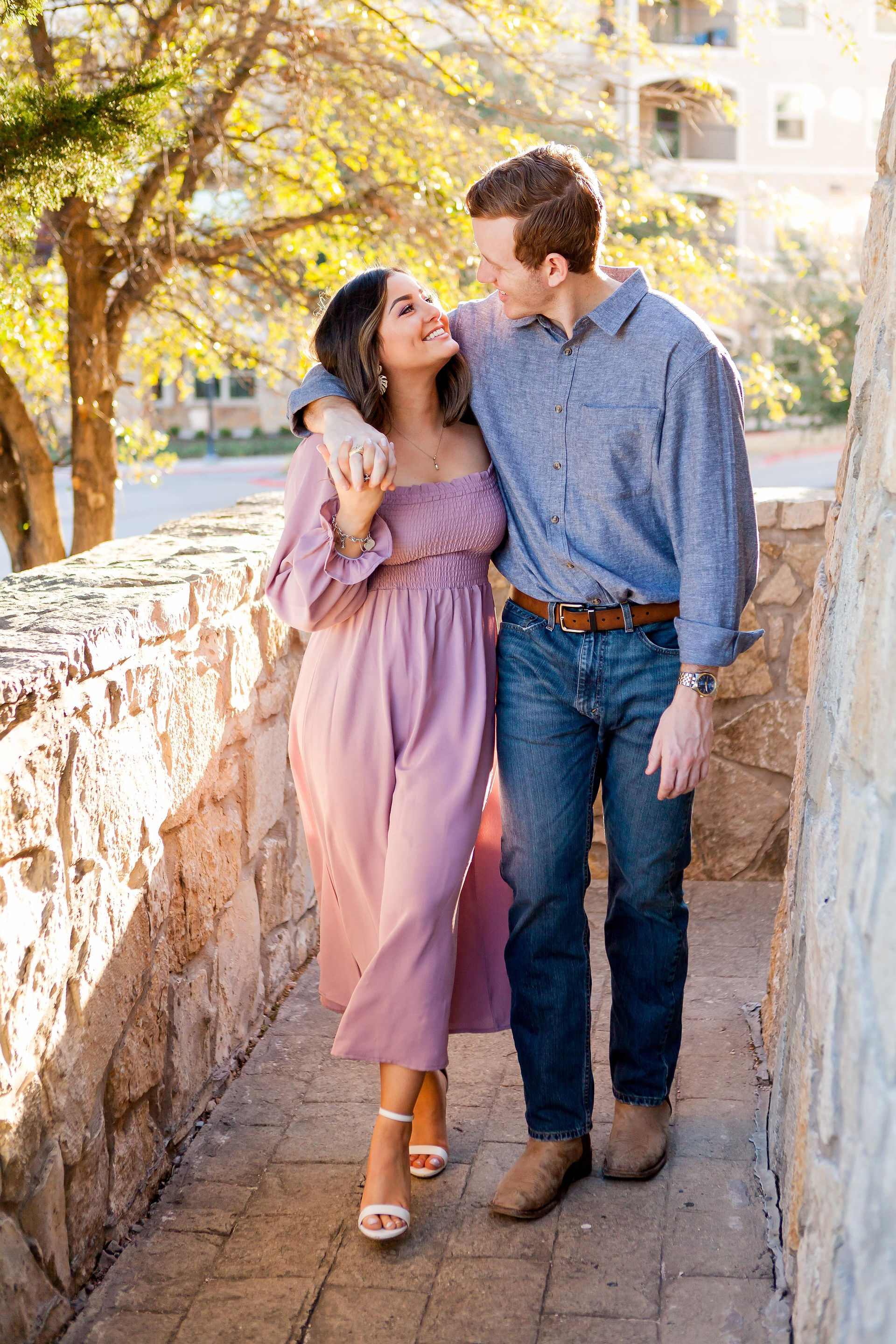 A newly engaged couple joyfully walk arm in arm along a narrow, rock-lined path as their picture is taken during their Dallas engagement photo session at the Adriatica Village in McKinney, TX. 
