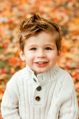 A boy smilingly gazes up at the camera while sitting in the fall leaves during his fall family photoshoot in Dallas, TX. Image captured by Photography of Sarah Mae.