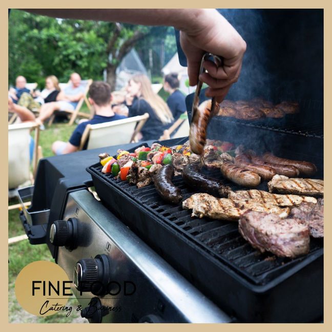 Grill w gronie najbliższych. Szef kuchni grilluje kiełbaski, kurczaka, steki i szaszłyki. Goście siedzą w tle na leżakach i rozmawiają. Catering live cooking zorganizowany jest w lesie na polance.