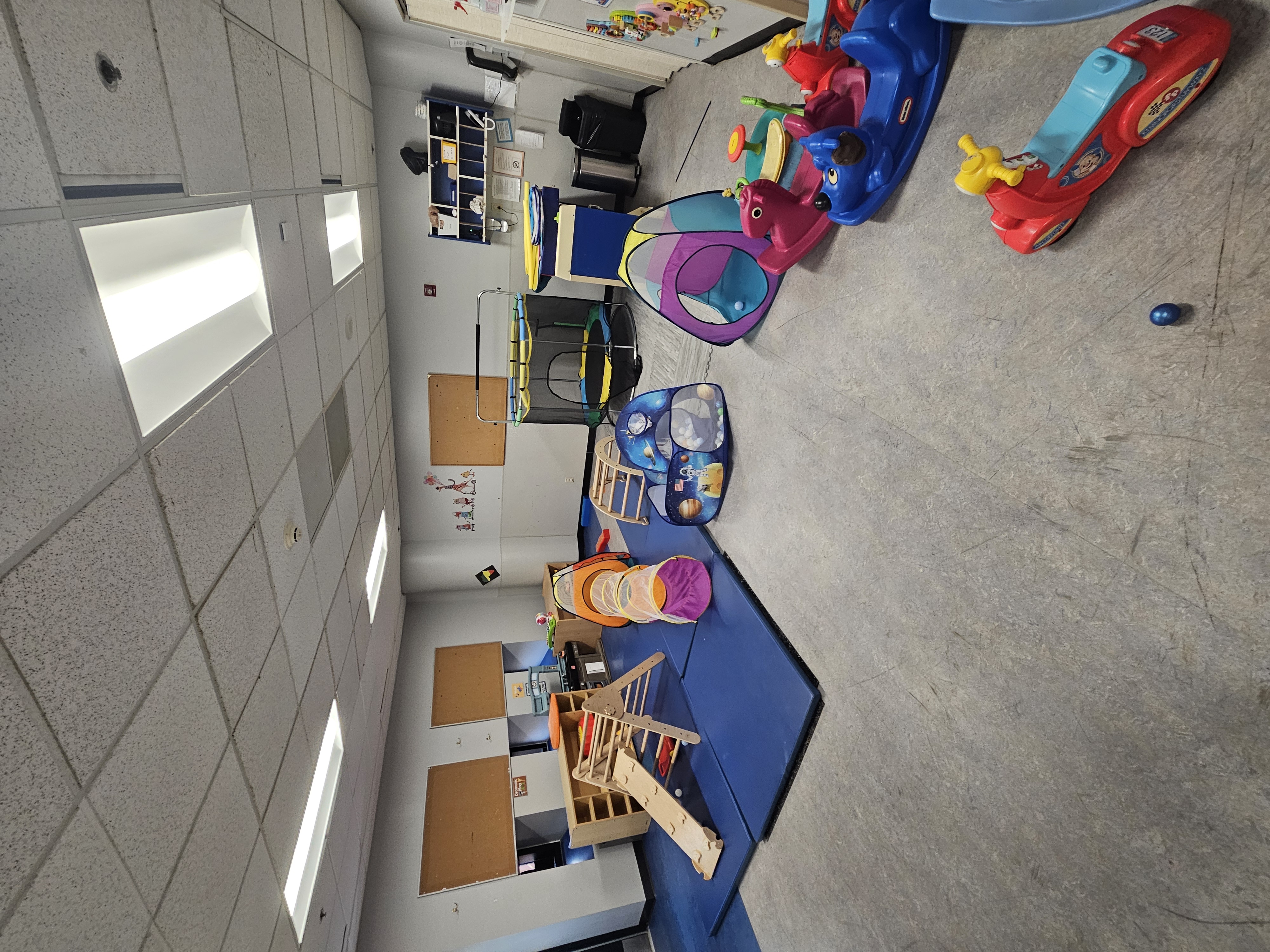 Daycare gross-motor room with blue mats, a wooden Pikler climbing ramp, colorful crawl tunnels, a small trampoline and plastic ride-on toys.