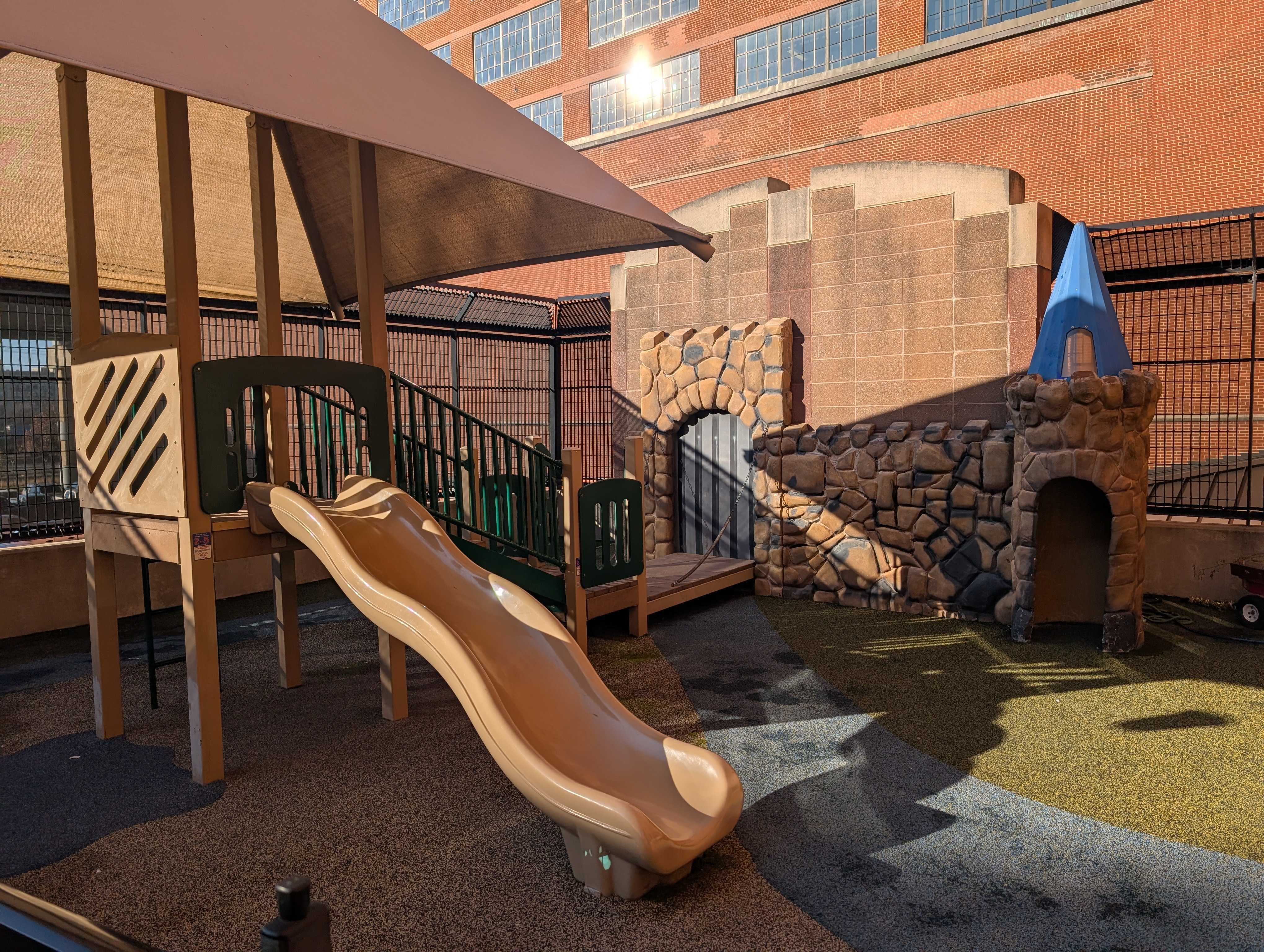 Childcare outdoor playground with a multi-level play structure, tan wavy slide, blue shade canopy, stone-look playhouse and green safety turf.