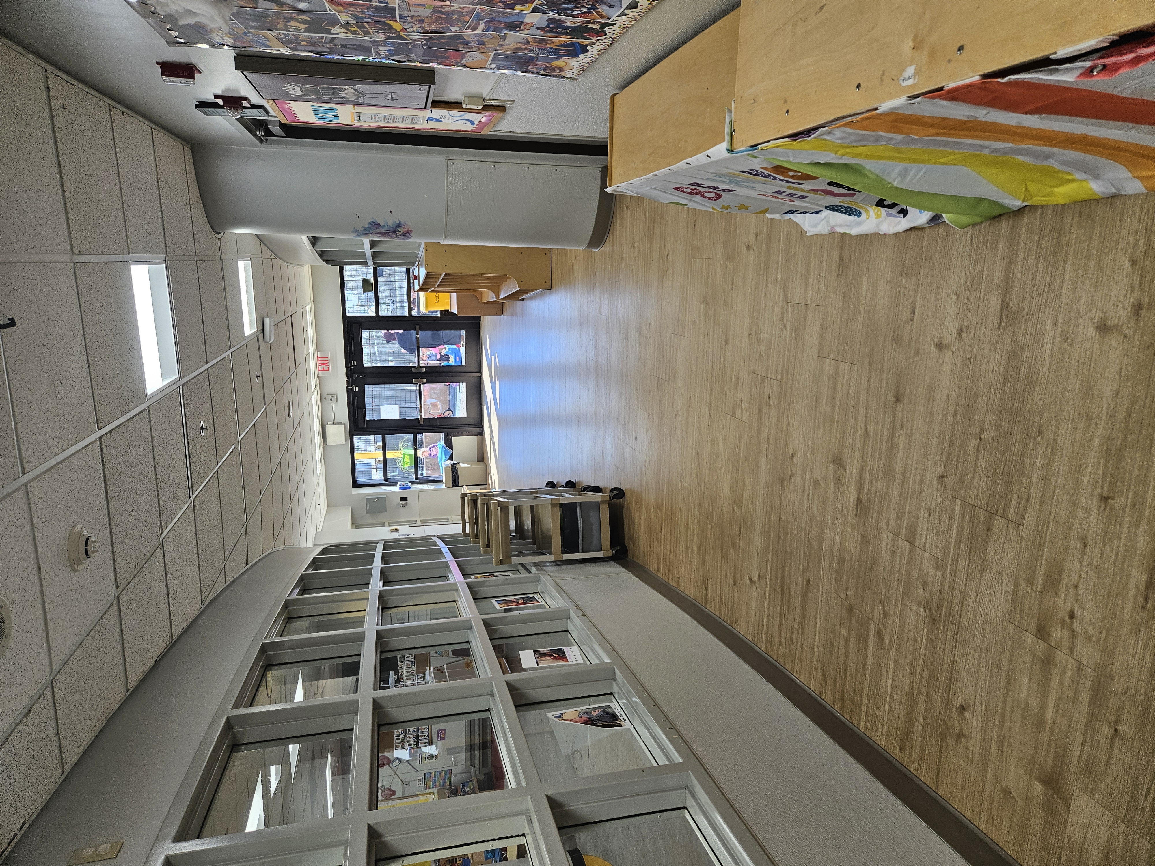 Childcare center hallway featuring a long row of interior windows into a toddler classroom, stacked mobile trays, wood-look flooring and glass double doors leading to an outdoor play yard