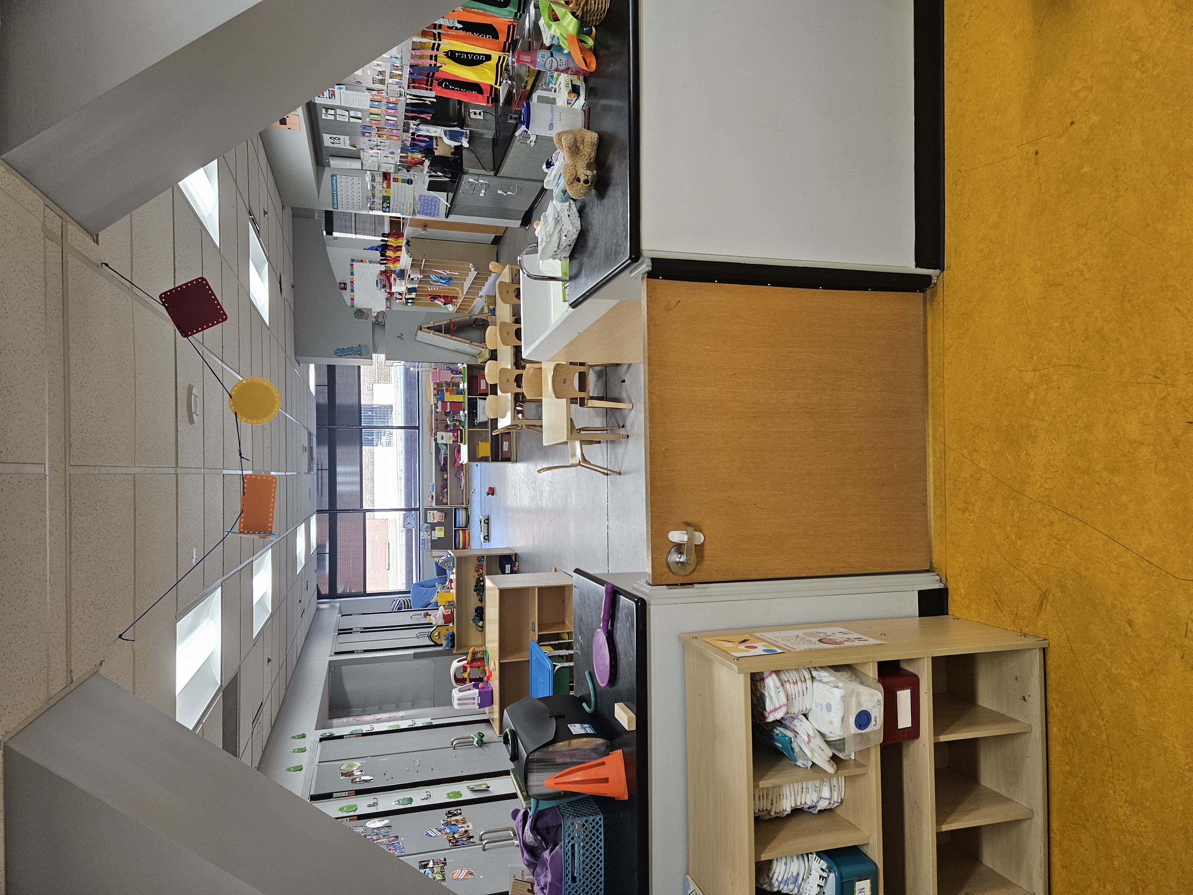 Bright toddler daycare classroom viewed through a child-height gate, featuring small wooden tables and chairs, low shelves of toys and diapers, a sink/counter area and activity centers under a large window.