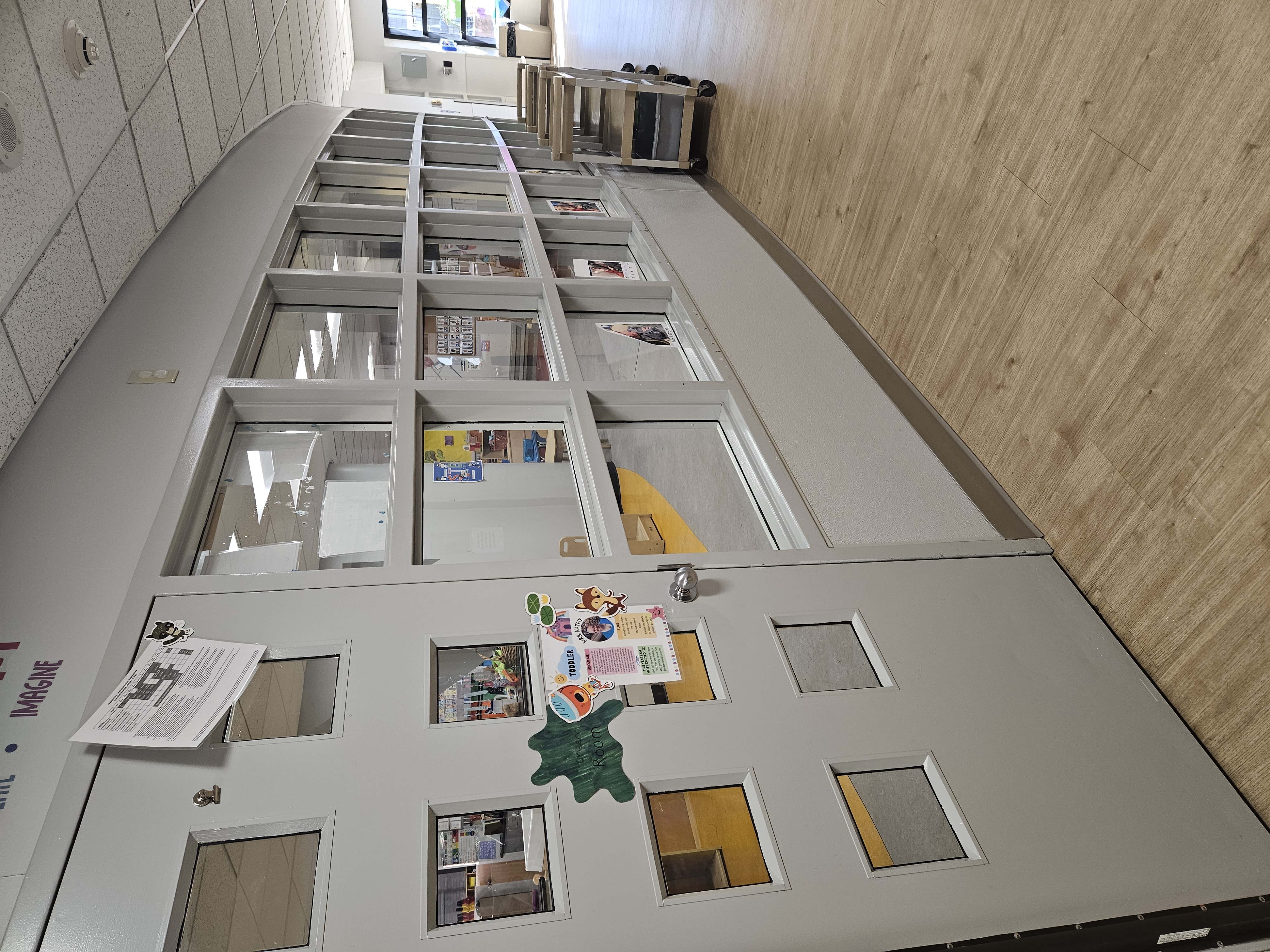 Daycare hallway showing the toddler classroom entrance with a gray multi-pane door, stickers, long interior windows, mobile supply carts and wood-look flooring.