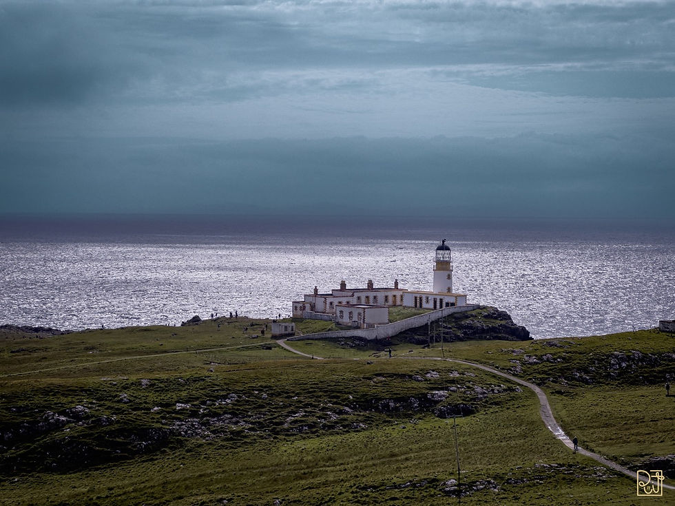 Un faro su una scogliera costiera sotto un cielo blu nuvoloso, con un terreno verdeggiante e frastagliato in primo piano. Il mare riflette una luce argentea.