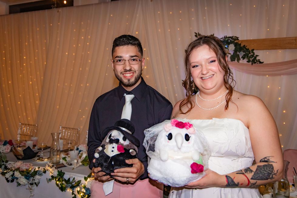 Couple smiles at each other holding their matching plush bride and groom dolls at the reception table