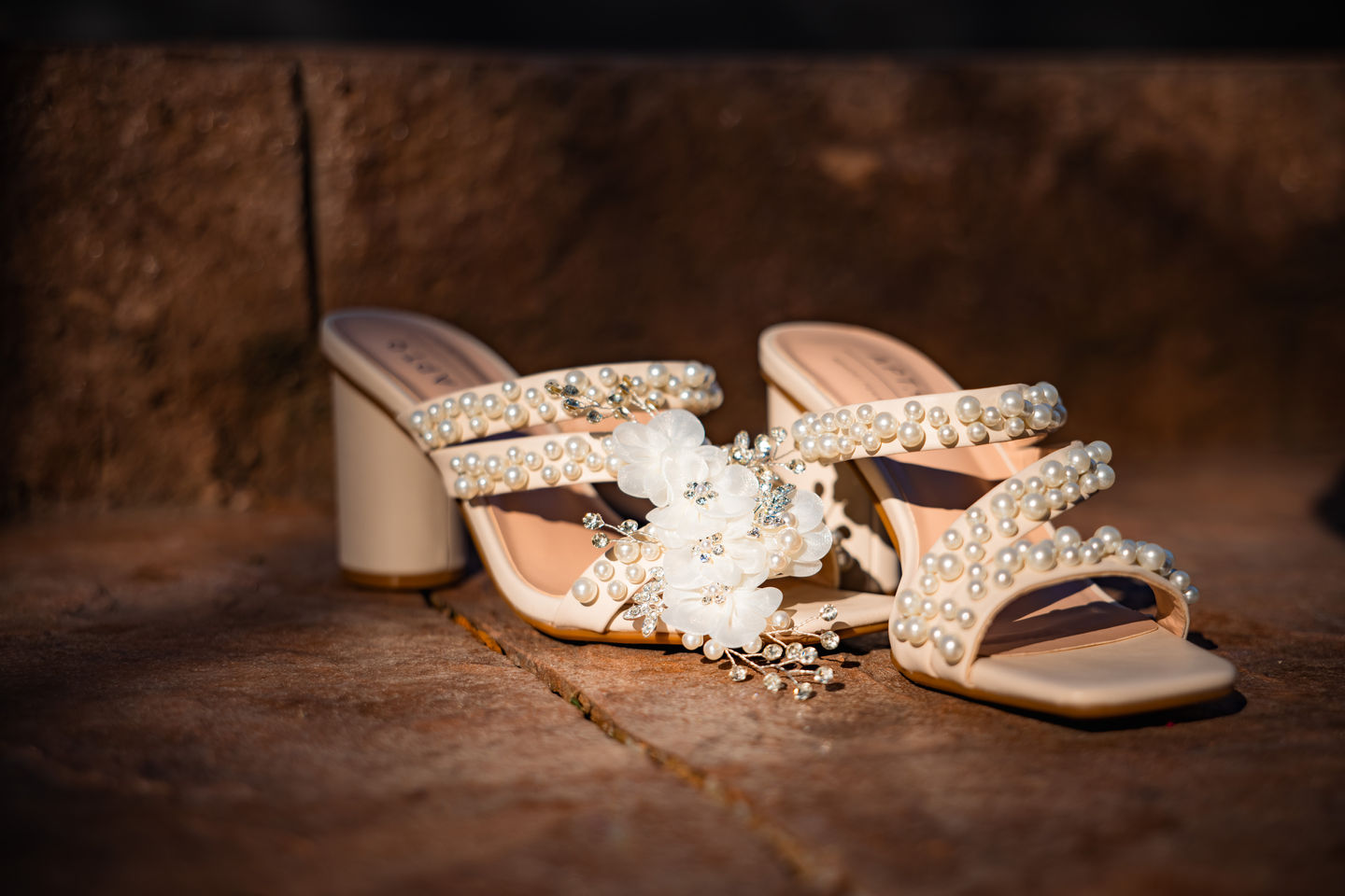 Pearl-embellished white bridal heels with floral details rest on a stone surface in soft natural light