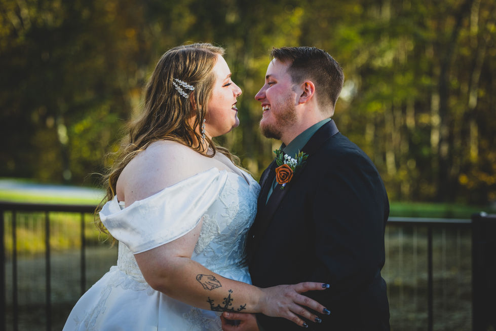 Bride and groom smile at each other near black railing with pond and autumn trees in background