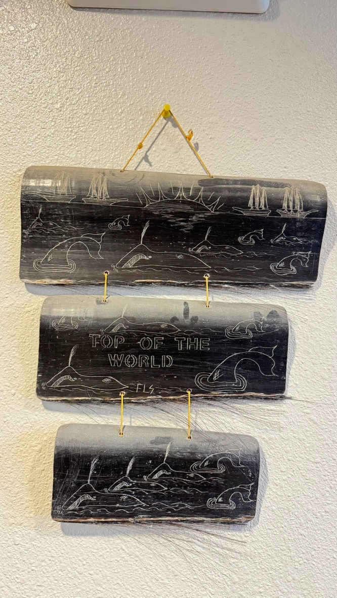“Top of the World.” A three-panel baleen scrimshaw, signed FLS, hung as a triptych: schooners standing off the horizon, whales spouting and sounding among small islands, and the inscription across the middle panel. The piece keeps in view a nineteenth-century Arctic that was, for a period, principally an American whaleship’s view of a coast the Iñupiat had already been living on for four thousand years. Photograph by John J. King II.