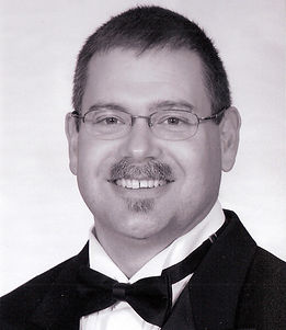 Headshot of Timothy Smith on a gray background, wearing a black suit and glasses.