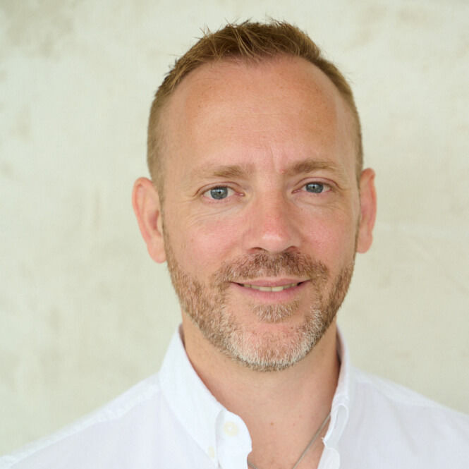 Smiling man with light beard in white shirt, set against a pale, plain background, conveying a friendly and relaxed mood.