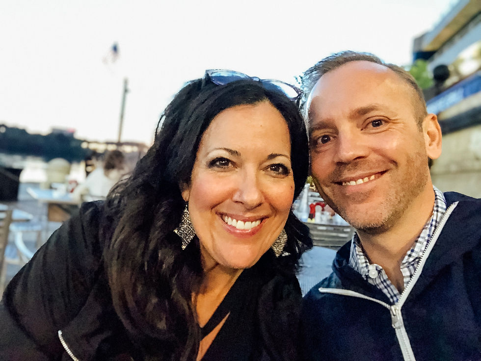 Smiling couple taking a selfie outdoors at a riverside café. Evening light, blurred tables, and an American flag in the background.
