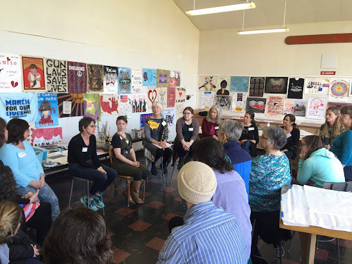 A diverse group of people sit in a circle in a room decorated with protest posters. They appear engaged in discussion.