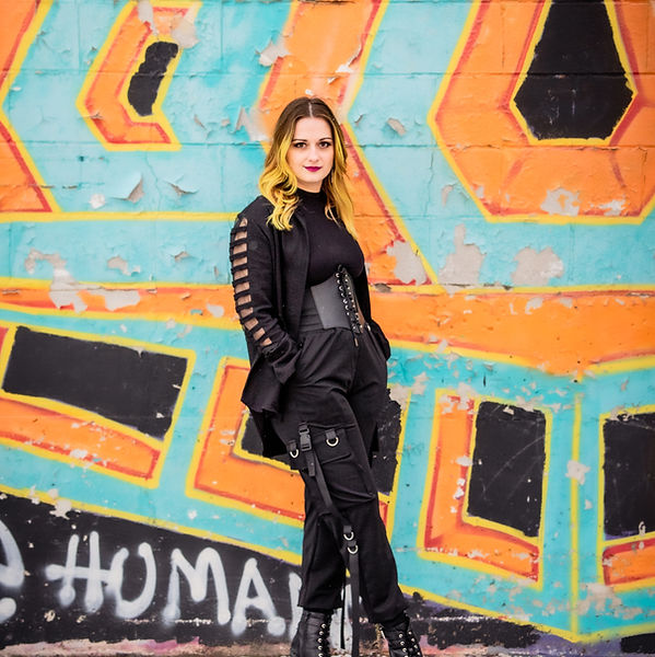 a white woman with mid length yellow hair, wearing a techwear outfit in all black, leans against a brightly graffiti painted wall