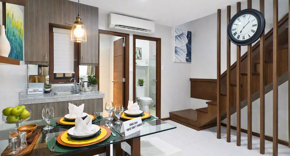 Modern dining and kitchen area with glass‑top table set for four, wooden cabinets, granite countertop, staircase with wall clock, and bright contemporary décor.