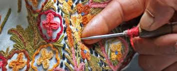 Close-up of hands doing intricate embroidery with a needle on colorful floral fabric, featuring red, yellow, and green threads.