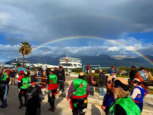 Colorful carnival celebration in Lixouri Kefalonia with parade costumes, waterfront views, ferry boat, mountains, and a rainbow during the 2026 carnival festivities.