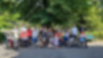 Eine Gruppe von 24 Menschen posiert lachend unter einem großen, grünen Baum. Einige Personen sitzen in Rollstühlen, andere stehen oder hocken daneben. Es ist ein sonniger Tag in einer grünen Parklandschaft.