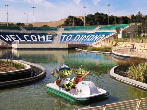 All About Aliyah mascots: Oleh and Olah the aliens, enjoying a paddle boat ride on the water in Dimona, Israel. Oleh takes a selfie while Olah waves, set against a park backdrop featuring stadium seating that spells out "Welcome to Dimona."