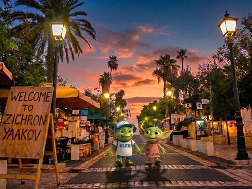 The All About Aliyah mascots, Oleh and Olah the aliens, holding hands while strolling along a charming cobblestone street at sunset in Zichron Yaakov. Oleh wears his signature blue Star of David cap and white "OLEH" shirt, while Olah wears a pink dress. A wooden sign on the left reads "WELCOME TO ZICHRON YAAKOV", framed by lit street lamps, outdoor cafe seating, and palm trees under a colorful evening sky.