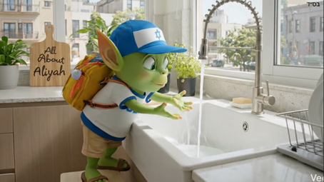 Oleh the Alien, the All About Aliyah mascot, wearing a blue Israel flag cap and a yellow backpack while standing on a stool to reach a running kitchen faucet in a bright, modern Israeli apartment. In the background, a wooden cutting board is visible with the All About Aliyah brand name next to a sink with a drying rack.