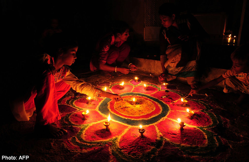 People light oil lamps around a vibrant rangoli pattern at night, creating a warm, festive atmosphere. Dark background enhances glow.