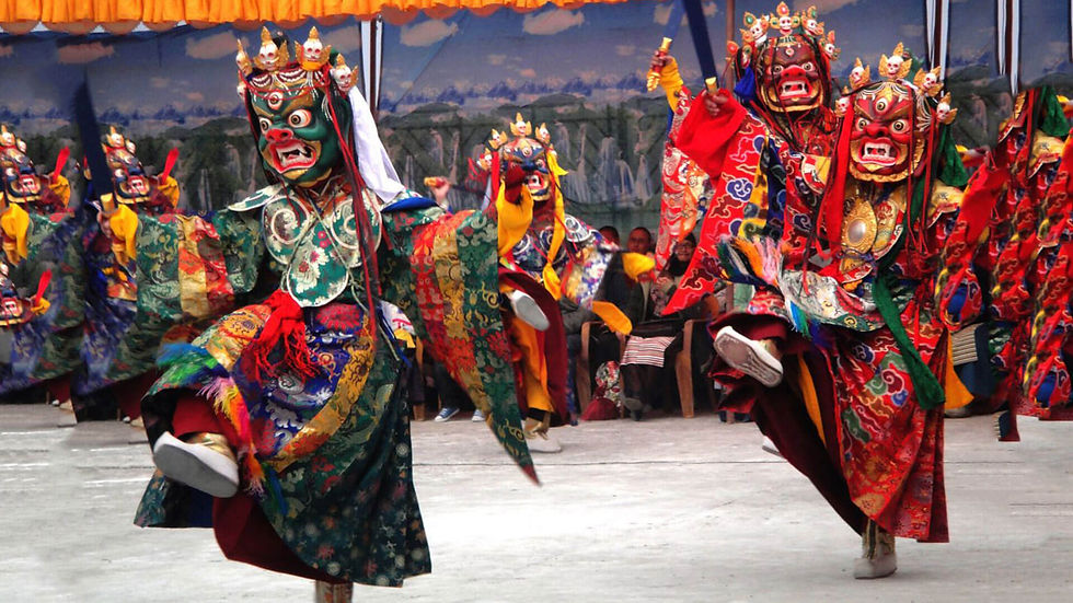Dancers in vibrant, ornate costumes and masks perform energetically in an outdoor setting, with a colorful backdrop and seated audience.