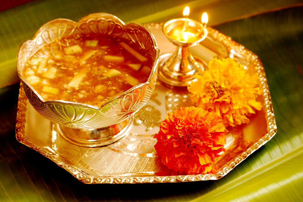 Golden tray with a bowl of liquid, surrounded by marigold flowers and a lit diya lamp, on a green leaf background. Warm festive ambiance.