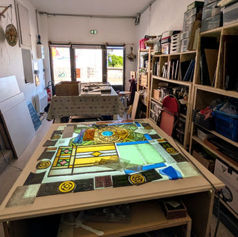 Atelier de vitrail d'Ingrid Redant à Malicorne-sur-Sarthe : grand vitrail à motifs floraux et géométriques colorés en cours d'assemblage sur une table lumineuse, étagères de verres et de matériaux en fond