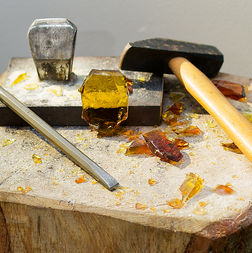 Outils du vitrailliste sur un billot de bois — marteline, tranchet et masse de fonte — entourés de blocs et d'éclats de verre ambré brut