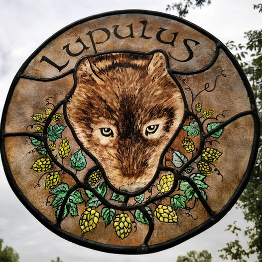 Vitrail rond « Lupulus » : tête de blaireau peinte à la grisaille, entourée de cônes de houblon jaunes et de feuilles vertes, cerné de plomb, photographié en transparence sur un ciel nuageux.