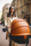 White woman wearing sunglasses and a beige trench coat, walking through the city while pushing a luxury brown stroller.