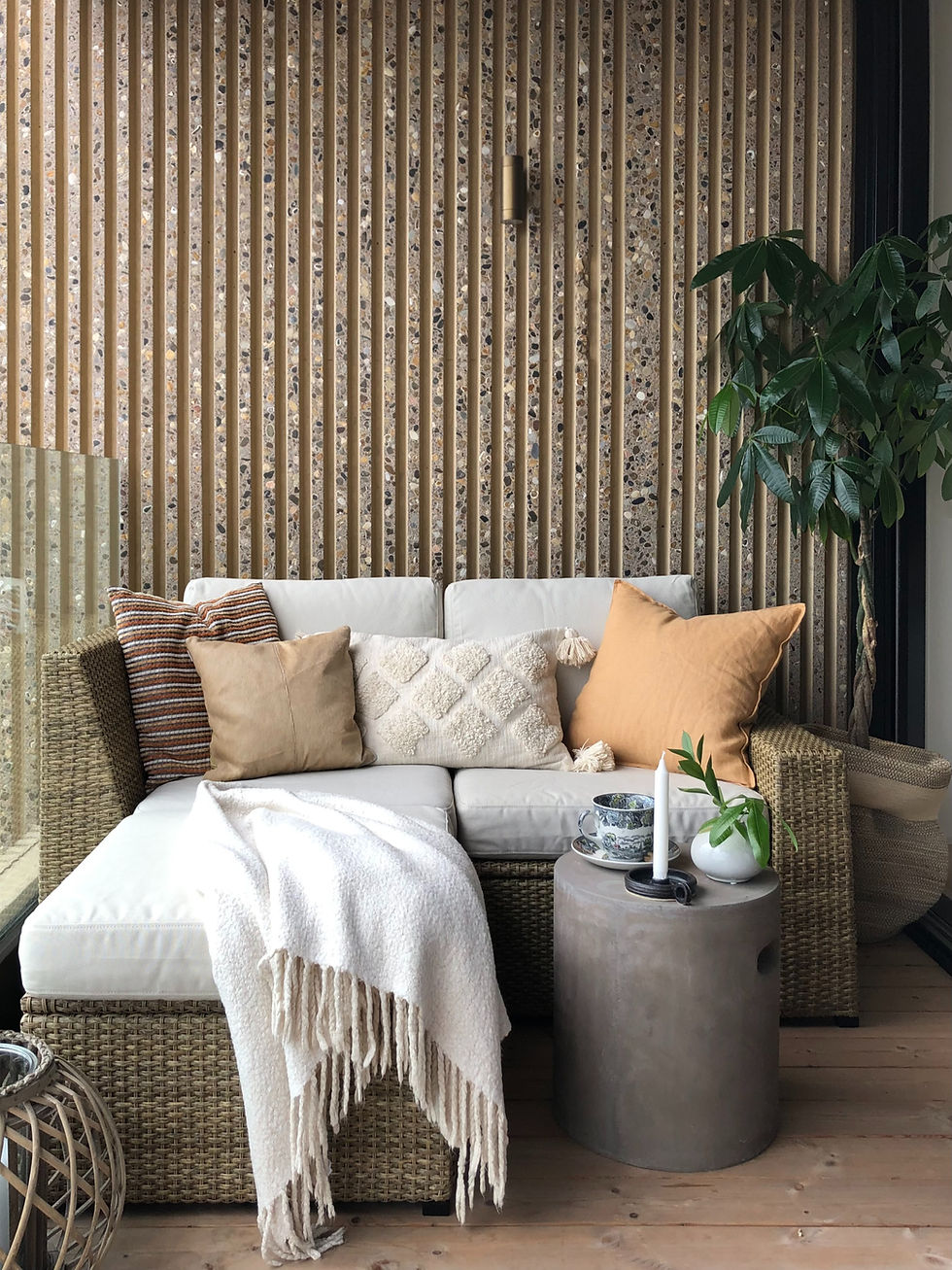 Balkong inredd med liten soffa i konstrotting, dynor och kuddar i ljusa toner och sidebord i betong