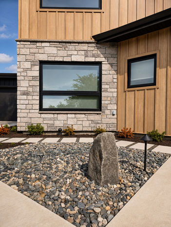 Front garden detail at Japavarian Haus in Richmond, showing stone cladding, warm vertical siding, black framed windows, gravel, planting, and sculptural stone.
