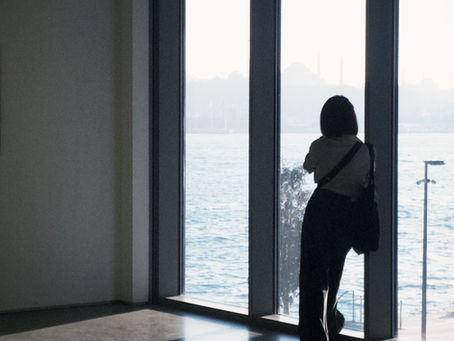 Woman standing by a large window overlooking the water, pausing in quiet reflection.
