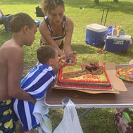 A boy wrapped in a towel blows out the candles on a birthday cake in a park