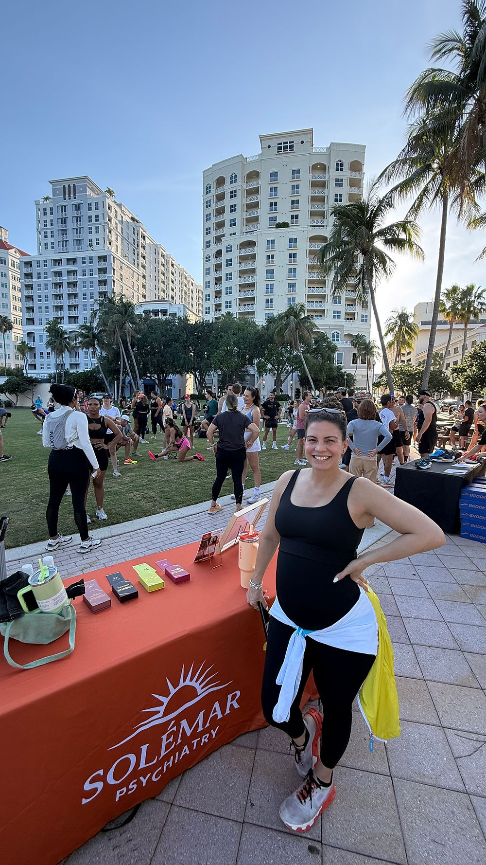 A smiling pregnant practitioner standing proudly next to a red SoléMar Psychiatry vendor table at a sunny outdoor fitness event in downtown West Palm Beach.