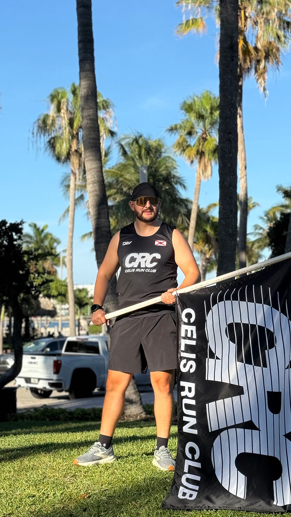 A runner holding a Celis Run Club flag between palm trees at a sunny West Palm Beach outdoor event, showcasing SoléMar Psychiatry's active community partnerships and support for local fitness groups.