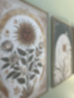 Close-up angled view of the leftmost botanical print showing an Art Nouveau-inspired sun face surrounded by roses and leaves in warm golds, creams, and greens within an ornate oval frame, with the adjacent dark-toned moonrise print partially visible.