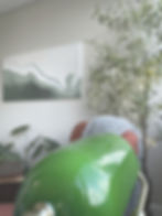 Detail shot of the green glass banker's lamp shade in the foreground, with the textured ocean wave painting, faux olive tree with fairy lights, monstera plant, and the coral armchair with gray throw visible in the soft-focus background.
