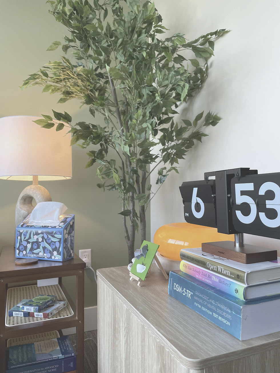 Detail shot of the side table and credenza featuring the DSM-5-TR, Dr. Julie Smith's books, a retro flip clock reading 6:53 PM, an amber glass mushroom lamp, a small green easel painting, and the blue floral tissue box beside the sculptural stone lamp.