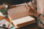 A person with a journal open in front of them on a wooden table. A cup of tea is next to the journal on the table.