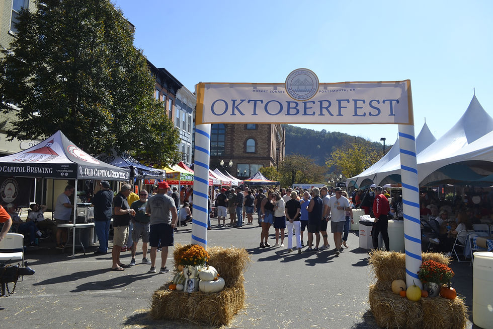 Oktoberfest event with lively crowd in sunny street, tents lining both sides, "OKTOBERFEST" banner overhead, and festive hay bales with pumpkins.
