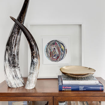 The image showcases an elegant home display with a blend of art and literature. A striking abstract sculpture, with smooth, intertwined forms in black and white, dominates the wooden console table. Behind it, a framed piece of circular abstract art adds a touch of color and complements the sculpture. On the table, a gold textured bowl sits atop a stack of hardcover books, including titles about Matisse and Picasso, indicating a taste for modern art. The lower shelf of the console displays a collection of books from various genres, and two framed black and white photographs add a personal touch, suggesting cherished memories. The arrangement is carefully curated, creating a sophisticated and cultured atmosphere.