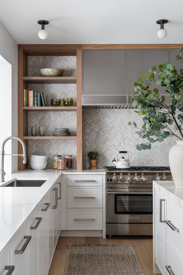 A stylish living room corner featuring a large fiddle leaf fig plant beside a white sofa adorned with cream and brown suede pillows. An abstract painting with greens, blues, and earth tones hangs on the wall. In the foreground, a black coffee table holds an assortment of books, including titles on architecture and art, as well as a decorative bowl. A rustic wooden horse sculpture adds a touch of whimsy to the setting.