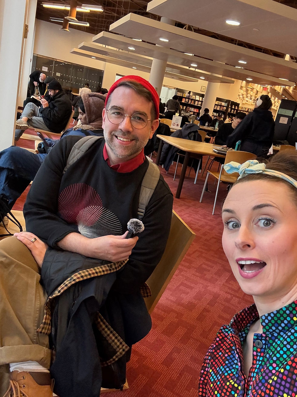 Smiling pair, Carl and Diana, take a selfie in a busy library lounge, with red carpet, bookshelves, and seated readers in the background, sitting on 'reverse-Z' seats in the Toronto Reference Library.